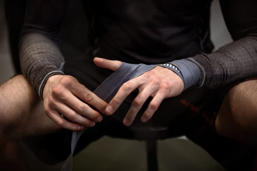 Male boxer sitting in dimly lit gym wrapping hands with protective boxing handwraps before training session to ensure safety and support, cropped image