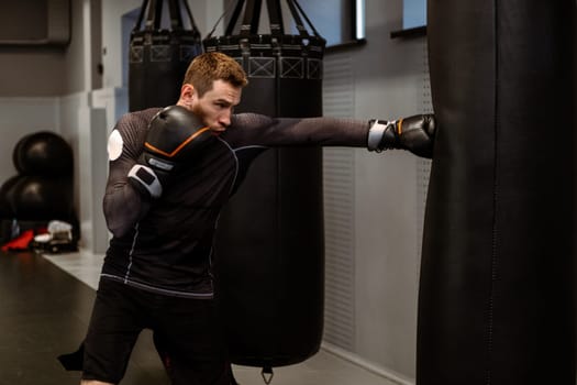 Young adult boxer focused on improving punch strength and power practicing on heavy bag while training alone in modern boxing gym. Sports motivation concept