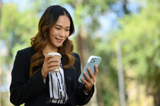 Cheerful professional financier using mobile phone, standing on background of green city park.