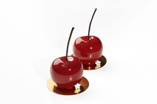 Two exquisite cherry-shaped desserts with mirror-like red glaze and delicate chocolate stems, presented on golden serving cardboards on white