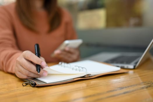 Woman using mobile phone and writing important things on notepad.