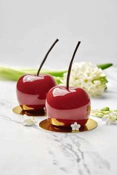 Two cherry-shaped pastries with lustrous red glaze and slender chocolate stems, displayed on gold cardboard cake bases accompanied by sprig of white flowers on marble top