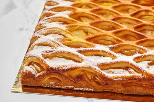 Close-up of golden lattice apple pie with tender custard and powdered sugar, freshly baked and displayed on golden cardboard on white surface