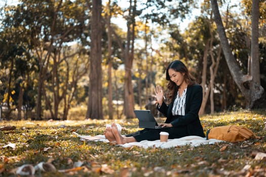 Carefree businesswoman sitting on grass in the autumn park and making a video call.