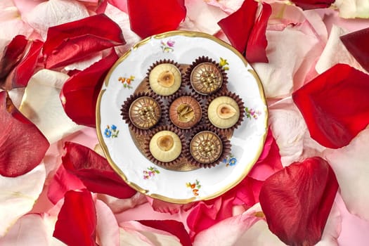 Elegant artisan chocolates decorated with hazelnuts, caramel crumbs and golden pearls arranged on vintage plate with floral design on background of colorful rose petals