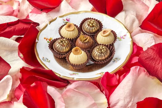Handmade candies from white, dark and milk chocolate decorated with nuts, caramel shavings and golden sugar pearls served on vintage plate amidst rose petals. Romantic sweet gift