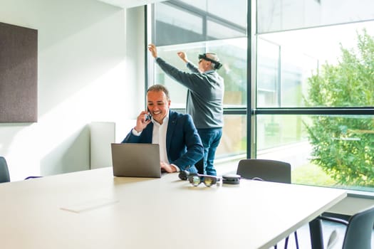 Businessman working using laptop while a colleague is using VR googles in the office