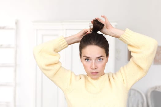 Serious emotionless woman in sweater looking at camera while setting hair in ponytail.