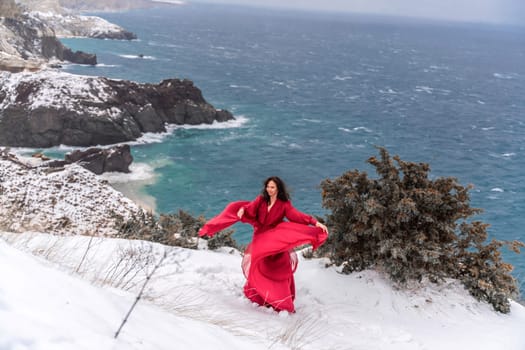 Woman red dress snow sea. Happy woman in a red dress in the snowy mountains by the emerald sea. The wind blows her clothes, posing against sea and snow background