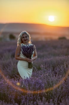 Blonde woman poses in lavender field at sunset. Happy woman in white dress holds lavender bouquet. Aromatherapy concept, lavender oil, photo session in lavender.