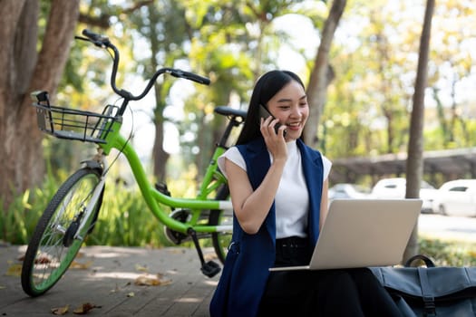 Asian businesswoman with bicycle using laptop and smartphone and sitting outside the office building. Woman commuting on bike go to work. Eco friendly vehicle, sustainable lifestyle concept.