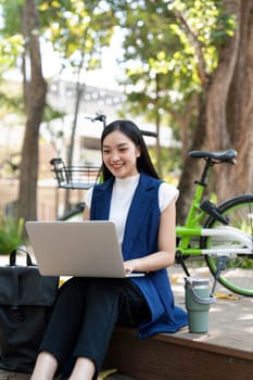Asian businesswoman with bicycle using laptop and sitting outside the office building. Woman commuting on bike go to work. Eco friendly vehicle, sustainable lifestyle concept.