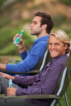 Camping, man and portrait with drinks for relax on grass in nature with happiness for holiday and vacation. People, smile and alcohol at campsite in woods for adventure, travel and bonding in forest.