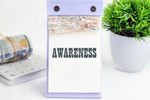 AWARENESS word writing on a desktop tear-off calendar on a white background, next to a calculator with a roll of banknotes with a flower out of focus in the background