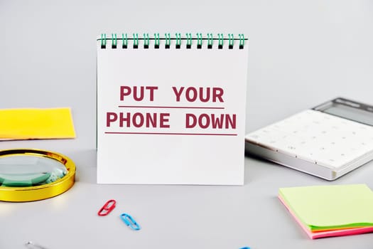 Put Your Phone Down text written on a blank sheet of notepad next to office supplies on a gray background. Business concept