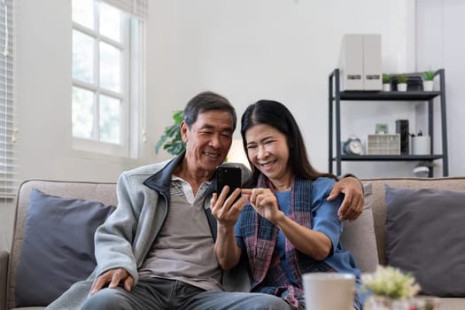 Happy elderly asian couple using smartphone sit on sofa doing ecommerce shopping online on website at home.