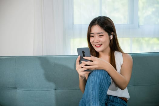 Happy young woman relaxed and messaging smartphone, Smiling Asian female sitting on sofa using mobile smart phone for shopping online at living room at home, Social media networks communication