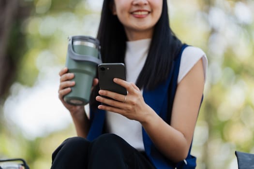 Smiling asian businesswoman hold reusable eco-friendly ecological cup and using mobile while sitting at park.