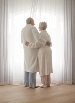 Senior couple, hug and relaxing by window in retirement, love and bonding in pajamas on holiday. Elderly people, back and embracing for affection in marriage, romance and morning routine at home.