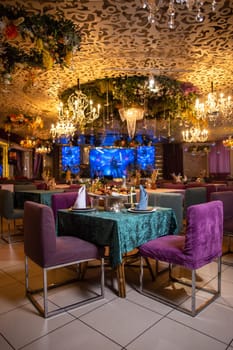 An elegant restaurant interior with purple chairs, green tablecloths, chandeliers, and decorative wall flowers creating a cozy atmosphere.