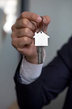 The image shows a persons hand holding a silver house key with a house-shaped keychain. The key is isolated on a white background.