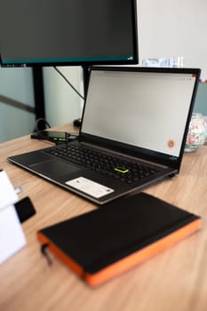 Open laptop with a blank screen, closed notebook with a pen on top, set on a light brown wood table in a cozy workspace setting.