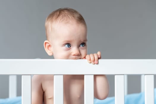 The happy baby, a toddler, is leisurely standing in the crib, looking over the bars with a smile on