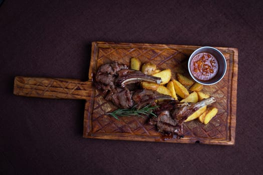 Two succulent lamb chops and crispy potato wedges served on a rustic wooden board with a side of flavorful dipping sauce.