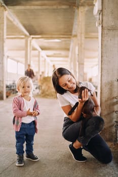 Little girl stands near her mother squatting down with a goatling in her arms. High quality photo