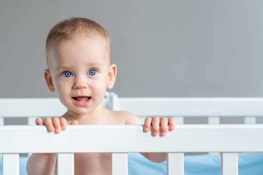 A girl with blue eyes stands in her crib with her mouth open in surprise.