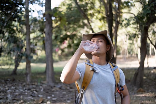 Asian male traveler carrying a large backpack drinks water from a bottle while resting during a hike..