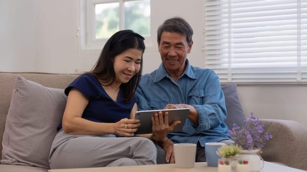 Cheerful Asian couple using network video call tablet sitting together on sofa at home Asian elderly husband sitting with elderly wife shopping online or video calling on tablet, relaxing..