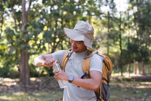 Asian male traveler carrying a large backpack drinks from a bottle while resting during a hiking trip..