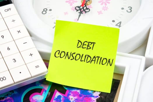 Debt consolidation. This is the process of obtaining a new loan to repay a number of existing debts. The text is written on a yellow sticker near the calculator and the clock