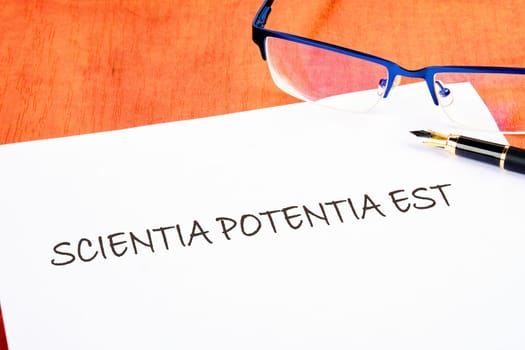 the phrase Scientia Potentia Est (Knowledge is power) written in Latin on a white piece of paper on the table next to the glasses