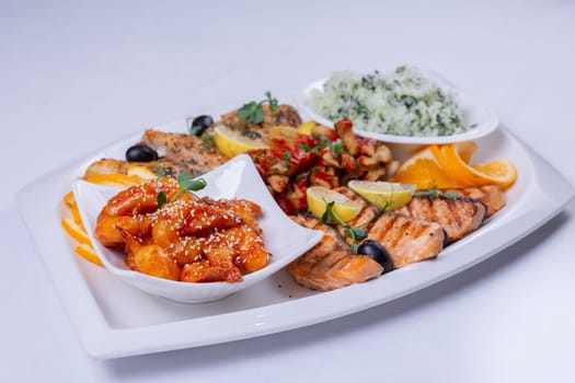 An assortment of grilled fish, sweet and sour chicken, and rice pilaf is artfully arranged on a white platter.