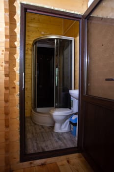 Wooden house bathroom with corner shower, toilet, sink. Glass door, wood walls floor. Cozy, modern, rustic design.