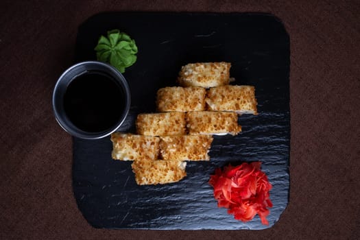 Deep-fried sushi roll on black stone plate with soy sauce, wasabi. Made with rice, seaweed, vegetables, shrimp tempura.