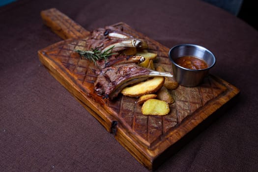 Two succulent lamb chops and crispy potato wedges served on a rustic wooden board with a side of flavorful dipping sauce.