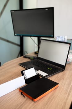 Open laptop with a blank screen, closed notebook with a pen on top, set on a light brown wood table in a cozy workspace setting.