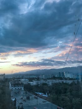 A stunning cityscape with mountains in the backdrop, surrounded by green trees under a blue sky with white clouds.