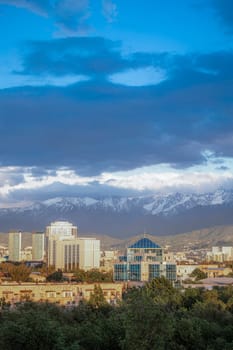 A stunning cityscape with mountains in the backdrop, surrounded by green trees under a blue sky with white clouds.