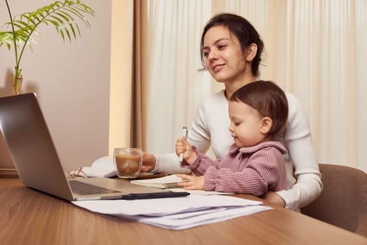 Cheerful pretty businesswoman working on laptop at home with her little child girl. mom spending time with her cute baby