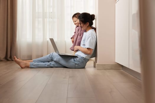 happy casual woman sitting on the floor and working on laptop with her little baby girl at home