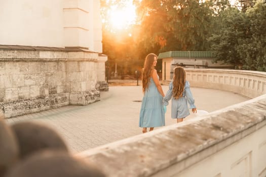 Daughter mother run holding hands. In blue dresses with flowing long hair, they hold balloons in their hands against the backdrop of a sunset and a white building