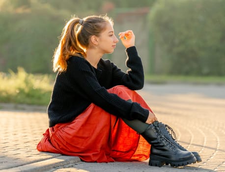 Teenage Girl Sits On City Sidewalk At Dawn In Backlight