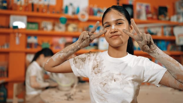 Happy caucasian girl pose at camera while diverse children modeling clay behind. Cute student wearing dirty shirt while looking at camera at workshop in art lesson. Blurring background. Edification.