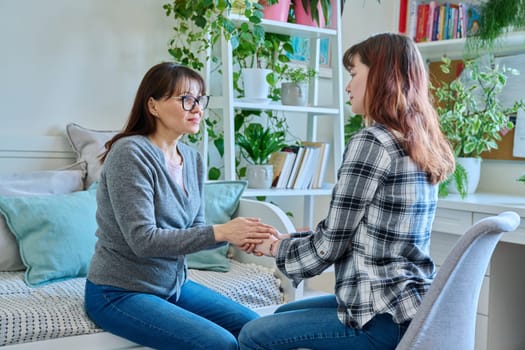 Talking mother and teenage daughter sitting together on couch at home, smiling holding hands. Family, communication, motherhood, friendship, relationship between parent and daughter 18-20 years old