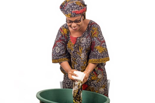 African woman standing above the laundry bowl washes clothes with a big piece of white soap isolated on white background.