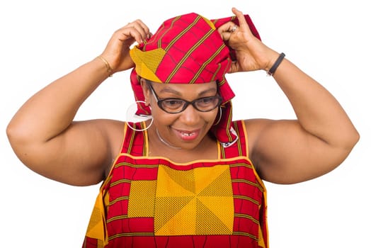 Close-up stylish smiling african mature woman in red african loincloth and glasses standing in studio and arranging her traditional headscarf on her head